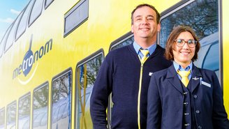 Zwei Personen in offizieller Bahnuniform stehen vor einem gelb-blauen Doppelstockzug mit dem Logo ‚metronom‘, aufgenommen am Bahnsteig bei Tageslicht.