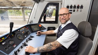 Lokführer sitzt im Führerstand eines modernen Zuges und bedient die Steuerungselemente, mit Blick auf die Instrumententafel und Gleise im Hintergrund.