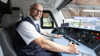 Lokführer sitzt im Führerstand eines Zuges und bedient die Steuerungselemente, Bahnhof Reichenbach im Hintergrund sichtbar.