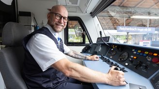 Lokführer sitzt im Führerstand eines Zuges und bedient die Steuerungselemente, Bahnhof Reichenbach im Hintergrund sichtbar.
