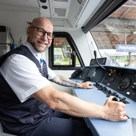 Lokführer sitzt im Führerstand eines Zuges und bedient die Steuerungselemente, Bahnhof Reichenbach im Hintergrund sichtbar.