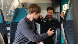 Ticketkontrolle im modernen Regionalzug, Fahrgast zeigt Fahrkarte, während Zugbegleiter ein mobiles Prüfgerät verwendet, Innenraum mit blauen Sitzen und Monitoren.