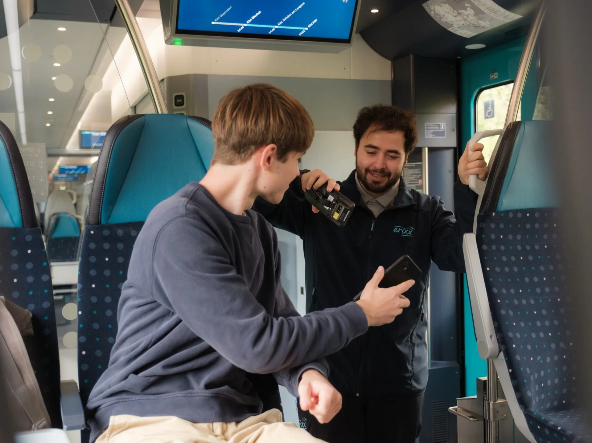 Ticketkontrolle im modernen Regionalzug, Fahrgast zeigt Fahrkarte, während Zugbegleiter ein mobiles Prüfgerät verwendet, Innenraum mit blauen Sitzen und Monitoren.