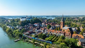 Regionalzug fährt entlang eines Flusses vor einer historischen Altstadt mit roten Dächern, Kirche und Schloss, umgeben von grüner Landschaft und blauem Himmel.