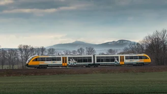 Moderner Regionalzug der ODEG fährt durch offene Landschaft mit grünen Feldern und kahlen Bäumen, Berge im Hintergrund unter bewölktem Himmel.