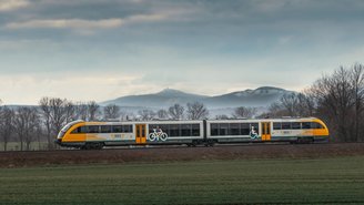Moderner Regionalzug der ODEG fährt durch offene Landschaft mit grünen Feldern und kahlen Bäumen, Berge im Hintergrund unter bewölktem Himmel.