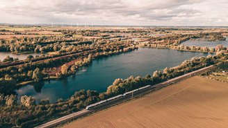 Regionalzug fährt entlang eines Sees durch eine weitläufige Landschaft mit Feldern, Bäumen und Wasserflächen unter bewölktem Himmel, aufgenommen aus der Luft.
