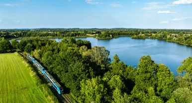 Regionalzug fährt durch grüne Landschaft entlang eines großen Sees unter blauem Himmel, umgeben von Wäldern und Wiesen.