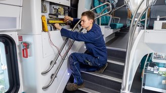 Techniker führt elektrische Wartungsarbeiten im Innenraum eines Doppelstockzugs durch, geöffnetes Schaltschrankpanel mit Kabeln und Messgerät sichtbar.