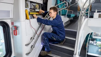 Techniker führt elektrische Wartungsarbeiten im Innenraum eines Doppelstockzugs durch, geöffnetes Schaltschrankpanel mit Kabeln und Messgerät sichtbar.