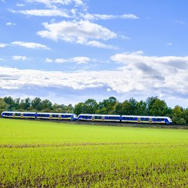 Blau-gelber Regionalzug fährt durch eine grüne Landschaft unter blauem Himmel mit weißen Wolken, umgeben von Feldern und Bäumen.