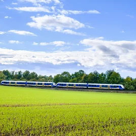 Blau-gelber Regionalzug fährt durch eine grüne Landschaft unter blauem Himmel mit weißen Wolken, umgeben von Feldern und Bäumen.