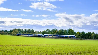 Blau-gelber Regionalzug fährt durch eine grüne Landschaft unter blauem Himmel mit weißen Wolken, umgeben von Feldern und Bäumen.