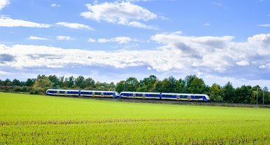 Blau-gelber Regionalzug fährt durch eine grüne Landschaft unter blauem Himmel mit weißen Wolken, umgeben von Feldern und Bäumen.