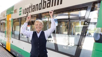 Person in Dienstkleidung steht vor einem grünen und weißen Zug mit der Aufschrift ‚vogtlandbahn‘ und zeigt mit beiden Händen nach oben.