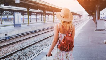 Reisende steht mit Koffer auf einem Bahnsteig, trägt Sommerkleid und Strohhut, bereit für die Zugfahrt.
