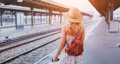 Reisende steht mit Koffer auf einem Bahnsteig, trägt Sommerkleid und Strohhut, bereit für die Zugfahrt.