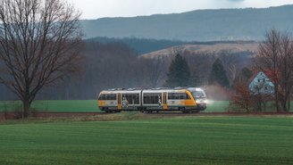 Moderner Regionalzug fährt durch grüne Felder vor bewaldeten Hügeln unter bewölktem Himmel