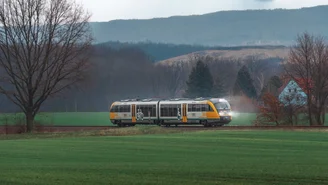 Moderner Regionalzug fährt durch grüne Felder vor bewaldeten Hügeln unter bewölktem Himmel