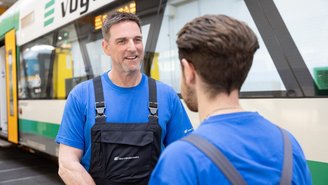 Zwei Mitarbeitende in blauen Arbeitsshirts und schwarzen Latzhosen stehen vor einem grünen und weißen Zug in einer Werkstatt und sprechen miteinander.