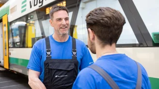 Zwei Mitarbeitende in blauen Arbeitsshirts und schwarzen Latzhosen stehen vor einem grünen und weißen Zug in einer Werkstatt und sprechen miteinander.