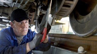 Mechaniker führt Wartungsarbeiten an Zugfahrwerk durch, Nahaufnahme von Werkzeug und Radkomponenten in einer Werkstattumgebung.