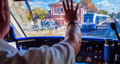 Blick aus dem Führerstand eines Zuges, während eine Person einem entgegenkommenden erixx‑Regionalzug an einem kleinen Bahnhof zuwinkt.