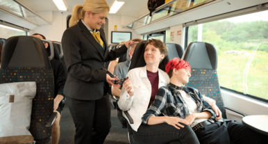 Zugbegleiterin überprüft Fahrkarten von Reisenden in einem modernen Regionalzug mit komfortablen Sitzen und großen Fenstern, während die Landschaft vorbeizieht.