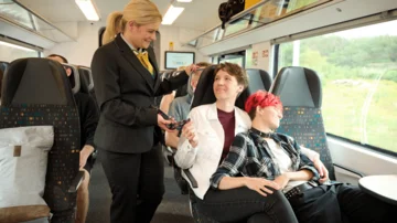 Zugbegleiterin überprüft Fahrkarten von Reisenden in einem modernen Regionalzug mit komfortablen Sitzen und großen Fenstern, während die Landschaft vorbeizieht.