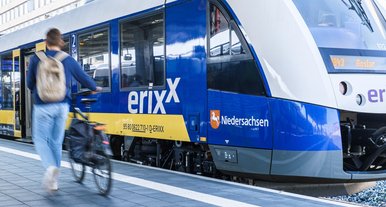 Blau-gelber Regionalzug der erixx GmbH steht am Bahnsteig, während eine Person mit Fahrrad vorbeigeht, Bahnhofsumgebung im Hintergrund.
