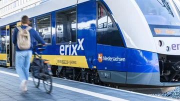 Blau-gelber Regionalzug der erixx GmbH steht am Bahnsteig, während eine Person mit Fahrrad vorbeigeht, Bahnhofsumgebung im Hintergrund.