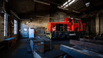 Rote Rangierlok steht in einer alten Werkstatthalle mit offenen Wartungsklappen, umgeben von Werkbänken und Metallteilen