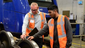 Zwei Personen in Warnwesten inspizieren Zugräder in einer Werkstatt vor einem blauen vlexx-Zug