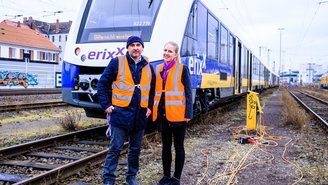 Zwei Personen in orangefarbenen Warnwesten stehen vor einem modernen erixx Regionalzug auf einem Gleisbereich mit Oberleitungen und Bahninfrastruktur im Hintergrund.