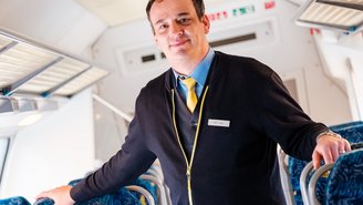 „Bahnmitarbeiter in offizieller Uniform steht im Innenraum eines modernen Regionalzugs, mit blauen Sitzreihen und heller Decke, gut sichtbare Details wie Notausgangsschilder und Gepäckablagen.