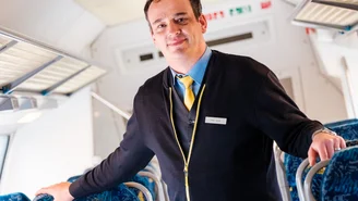 „Bahnmitarbeiter in offizieller Uniform steht im Innenraum eines modernen Regionalzugs, mit blauen Sitzreihen und heller Decke, gut sichtbare Details wie Notausgangsschilder und Gepäckablagen.