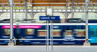 Regionalzug fährt durch den überdachten Bahnsteig des Lübeck Hauptbahnhofs, unscharf in Bewegung, mit sichtbarem Stationsschild und Stahlkonstruktion im Hintergrund.