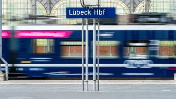 Regionalzug fährt durch den überdachten Bahnsteig des Lübeck Hauptbahnhofs, unscharf in Bewegung, mit sichtbarem Stationsschild und Stahlkonstruktion im Hintergrund.