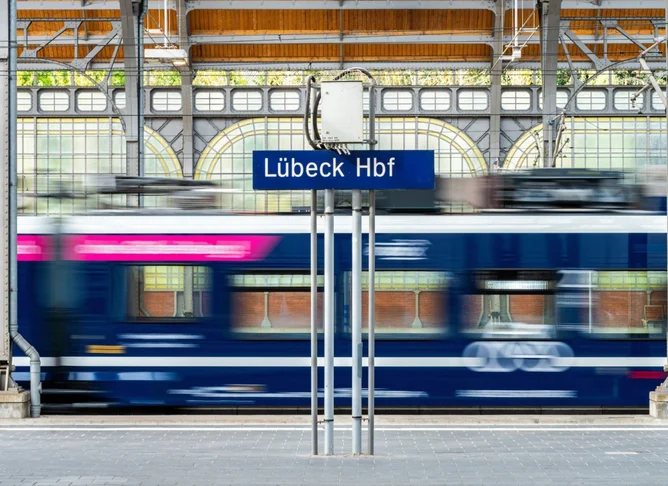 Regionalzug fährt durch den überdachten Bahnsteig des Lübeck Hauptbahnhofs, unscharf in Bewegung, mit sichtbarem Stationsschild und Stahlkonstruktion im Hintergrund.
