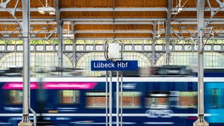 Regionalzug fährt durch den überdachten Bahnsteig des Lübeck Hauptbahnhofs, unscharf in Bewegung, mit sichtbarem Stationsschild und Stahlkonstruktion im Hintergrund.