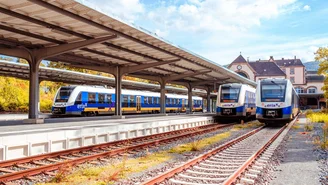 Drei blau-gelbe Regionalzüge stehen an einem überdachten Bahnsteig eines Bahnhofs mit historischem Gebäude und Gleisanlagen unter teils bewölktem Himmel.