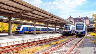 Drei blau-gelbe Regionalzüge stehen an einem überdachten Bahnsteig eines Bahnhofs mit historischem Gebäude und Gleisanlagen unter teils bewölktem Himmel.