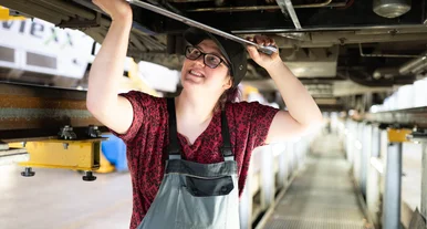 Mitarbeiterin führt Wartungsarbeiten unter einem Zugfahrzeug in einer Werkstatt durch, mit Werkzeug in der Hand.