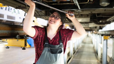 Mitarbeiterin führt Wartungsarbeiten unter einem Zugfahrzeug in einer Werkstatt durch, mit Werkzeug in der Hand.