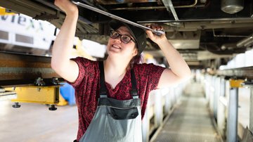 Mitarbeiterin führt Wartungsarbeiten unter einem Zugfahrzeug in einer Werkstatt durch, mit Werkzeug in der Hand.
