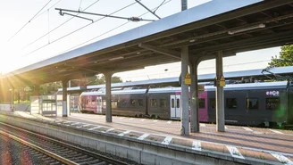 Moderner Regionalzug am sonnigen Bahnhof mit überdachtem Bahnsteig und Oberleitungen, Schienen im Vordergrund, klare Sommerbeleuchtung.