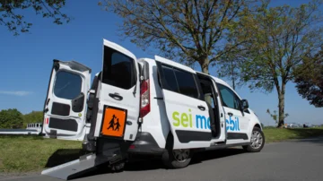 Weißer Kleinbus mit der Aufschrift ‚sei mobil‘ steht auf einer Straße, geöffnete Hecktüren mit ausfahrbarer Rampe für barrierefreien Zugang.