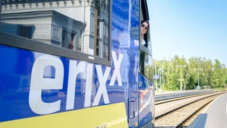 Blau-gelber Regionalzug der erixx GmbH steht am Bahnsteig, mit geöffnetem Fenster und Blick auf Gleise, Bahnsteig und grüne Bäume im Hintergrund.