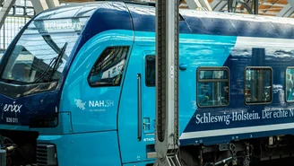 Blau-türkiser Regionalzug mit der Aufschrift ‚Schleswig-Holstein. Der echte Norden.‘ steht im überdachten Bahnhof, sichtbare Stahlträger und Oberleitungen im Hintergrund.