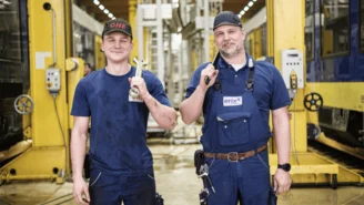 Zwei Personen in Arbeitskleidung stehen in einer Werkstatthalle zwischen gelb-blauen Zügen und halten Werkzeuge in der Hand